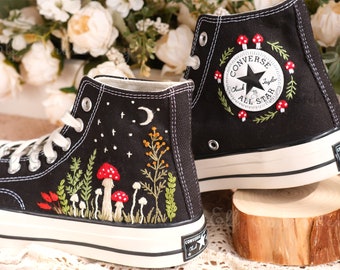 May include: A pair of black high-top Converse sneakers with embroidered designs. One shoe features a night sky with a crescent moon and stars, while the other has a floral wreath with red mushrooms around the Converse All Star logo.
