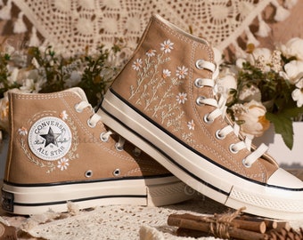 Puede incluir: Un par de zapatillas de lona de caña alta de color marrón con suelas blancas y cordones blancos. Las zapatillas están bordadas con flores blancas y hojas verdes. La zapatilla izquierda tiene el logotipo de Converse All Star bordado en el lateral.