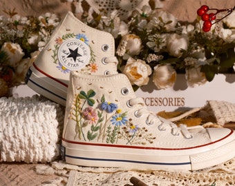 May include: White high-top Converse sneakers with embroidered flowers, leaves, and bees. The sneakers have a red stripe around the sole and the Converse All Star logo is visible on the side.
