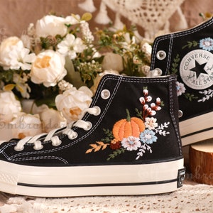 May include: Pair of black high-top sneakers with white soles and laces. The sneakers are adorned with colorful floral embroidery, including an orange pumpkin and various flowers. The Converse All Star logo is visible on the side.