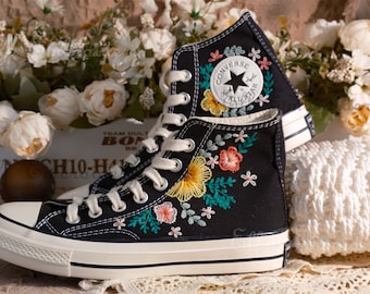 May include: Black high-top canvas sneakers with white laces and a white rubber sole. The sneakers are embroidered with colorful flowers and leaves. The Converse All Star logo is visible on the side of the shoe.