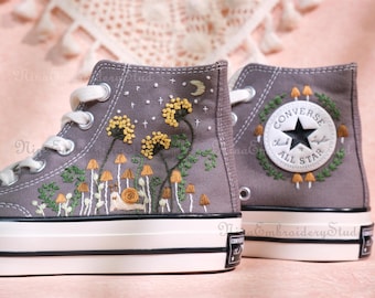 May include: Gray high-top sneakers with custom embroidery. The design features yellow flowers, mushrooms, a snail, and a crescent moon. The heel displays the Converse All Star logo. The shoes have white laces and soles.