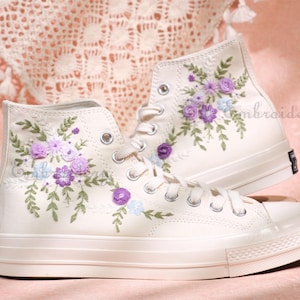 May include: Off-white high-top sneakers with embroidered floral designs in purple, lavender, and light blue. The floral embroidery is on the outer sides of the shoes, with green leaves. The sneakers have white laces and a white rubber sole.
