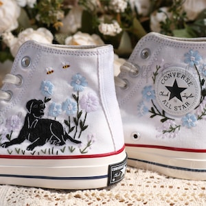 Könnte beinhalten: Weiße High-Top-Sneaker mit gestickten Blumen- und Hunde-Designs. Die Sneaker zeigen eine schwarze Hundesilhouette, blaue und lila Blumen und zwei Bienen. Das Converse-Logo ist sichtbar.