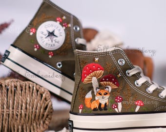 May include: A pair of olive green high-top canvas sneakers with embroidered details. The sneakers feature a fox, mushrooms, and bees. The left sneaker has a Converse All Star logo.