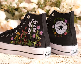 May include: Black high-top Converse sneakers with embroidered floral designs. One shoe features a floral wreath around the Converse All Star logo.
