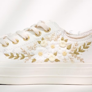 May include: White canvas sneakers with intricate floral embroidery. The design features cream-colored daisies and leaves with gold accents. The shoe has cream-colored laces and gold eyelets.