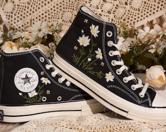 Puede incluir: Zapatillas Converse negras de caña alta con suelas blancas y un diseño bordado de margaritas y abejas. El logotipo de Converse All Star es visible en el lateral de la zapatilla.
