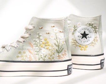 Könnte beinhalten: Hohe Canvas-Sneaker in einem sanften Grünton, verziert mit zarter Blumenstickerei. Das Design zeigt weiße Gänseblümchen, gelbe Blüten und kleine Bienen. Das Converse All Star-Logo ist auf der Rückseite zu sehen.
