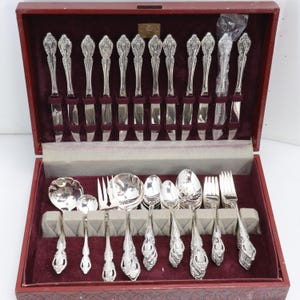Può includere: Un set di posate argentate presentato in una custodia in legno rosso scuro con interno in velluto. Il set comprende vari coltelli, forchette e cucchiai, ognuno caratterizzato da un design del manico ornato e in rilievo. Un coltello è avvolto in plastica.