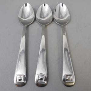 Może przedstawiać: Trzy błyszczące srebrne łyżki z kwadratowym wzorem jabłka u podstawy uchwytu. Łyżki mają klasyczny, elegancki design, idealny do serwowania. Łyżki wykonane są z odblaskowego metalu.