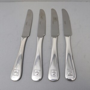 Może przedstawiać: Cztery srebrne noże do masła z zakrzywionymi ostrzami i zaokrąglonymi uchwytami. Każdy uchwyt ma kwadratowy emblemat z wzorem. Noże ułożone są na jasnoszarej powierzchni.