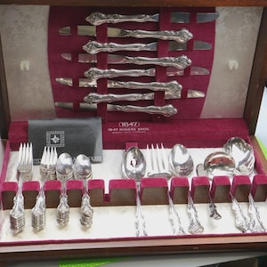 May include: A vintage silver cutlery set presented in a wooden case. The set includes knives, forks, spoons, and serving utensils, all with decorative handles. The case interior is lined with red velvet. The text "(1847) 1847 ROGERS BROS." is visible.