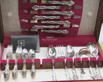 May include: A vintage silverware set in a wooden case. The set includes knives, forks, spoons, and serving utensils, all with ornate handles. The interior of the case is lined with red velvet. The text "(1847) 1847 ROGERS BROS." is visible.