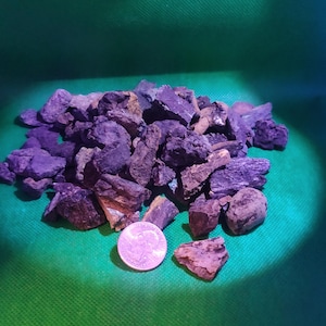 May include: A pile of dark purple and brown rocks with a quarter coin on a green background. The rocks vary in size and shape, with rough, textured surfaces. The coin is silver and shows a profile of a person.