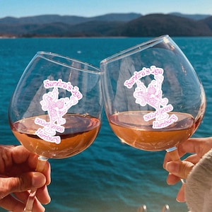 May include: Two wine glasses clinked together, filled with a rose-colored liquid. Each glass has a pink logo with text that reads "Sandra's Sea" and "30.03.2026 Hamburg". The background features a lake and mountains under a blue sky.
