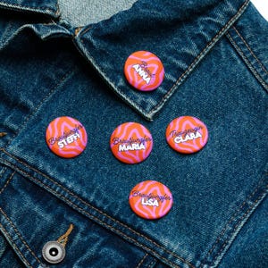 May include: A collection of orange and pink swirl-patterned buttons with names in white and purple text. The buttons are pinned to a blue denim jacket. The text on the buttons includes names like "Anna", "Steffi", "Maria", "Clara", and "Lisa".