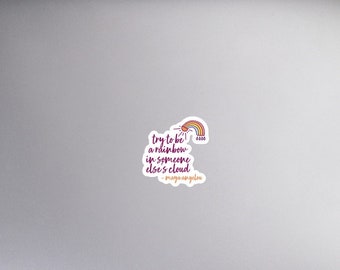May include: A laptop computer with a sticker that says "Try to be a rainbow in someone else's cloud." The sticker features a rainbow with a cloud and a sun.