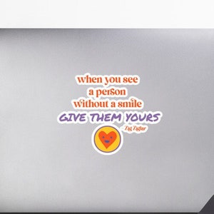 Puede incluir: Una pegatina con un diseño morado, naranja y amarillo. La pegatina presenta un corazón con una cara sonriente y el texto "when you see a person without a smile GIVE THEM YOURS" con las palabras "Fat Vader" debajo del corazón.
