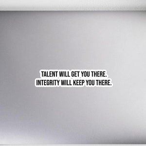 Op de afbeelding: Een witte sticker op een laptop met de tekst "TALENT WILL GET YOU THERE. INTEGRITY WILL KEEP YOU THERE." De laptop staat op een wit oppervlak met een notitieboek, potloden, een koptelefoon, een muis en een plant.