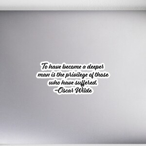 以下が含まれることがあります： 白いラップトップに、黒と白のステッカーが貼ってあります。ステッカーには、「より深い人間になることは、苦しんだ者の特権である。 -オスカー・ワイルド」と書いてあります。