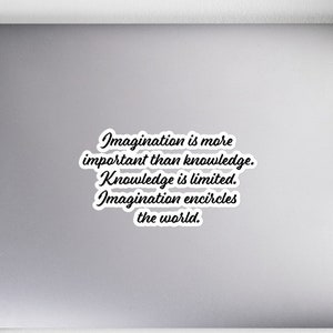 Pode incluir: Um adesivo branco em um laptop com a citação "A imaginação é mais importante que o conhecimento. O conhecimento é limitado. A imaginação envolve o mundo" em escrita preta. O laptop está em uma superfície branca.