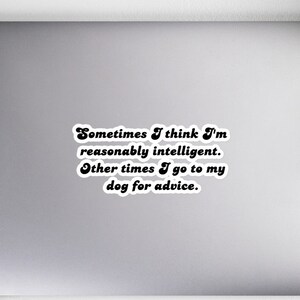 May include: A white sticker with black text that reads, "Sometimes I think I'm reasonably intelligent. Other times I go to my dog for advice." The sticker is on a gray laptop, with a notebook, pen, headphones, and a plant in the background.