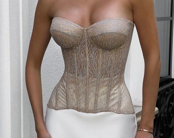 May include: A strapless corset with a beige and silver glitter pattern. The corset has a boned structure and a sweetheart neckline.
