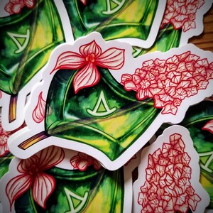 Könnte beinhalten: Eine Sammlung von Aufklebern mit einem grün-gelben Design und einem weißen Rand. Jeder Aufkleber hat ein stilisiertes rotes Blumenelement und ein weißes Symbol. Die Aufkleber sind auf einer Holzoberfläche verstreut.