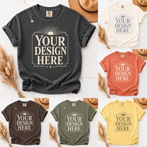 May include: A collection of six short-sleeved t-shirts in various colors, including charcoal, cream, orange, brown, olive green, and yellow. Each shirt features the text "YOUR DESIGN HERE" within a decorative frame, with a sunburst design above the text.