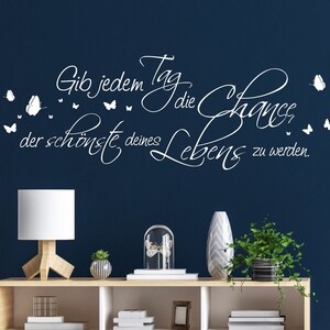 Può includere: Adesivo da parete bianco con il testo "Gib jedem Tag die Chance der schönste deines Lebens zu werden." L'adesivo presenta un design a farfalla stilizzato. L'adesivo è esposto su una parete blu scuro sopra un'unità a scaffale in legno con cassetti bianchi. L'unità a scaffale è decorata con una lampada bianca, una campana di vetro con una farfalla all'interno, una piccola pianta in vaso e un vaso con un motivo geometrico.