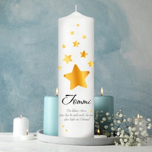 Peut inclure: Une bougie pilier blanche ornée d'une grande étoile dorée et de petites étoiles dorées. Le nom "Tommi" est écrit en script noir en dessous, avec un court message. Deux bougies bleues et une bougie blanche sont allumées à proximité.