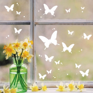 Könnte beinhalten: Eine Vase mit gelben Narzissen steht auf einem Fensterbrett mit Blick auf eine weiße, gefrostete Fensterscheibe, die mit weißen Schmetterlingsaufklebern verziert ist.