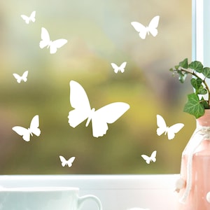 Può includere: Stili di finestra a forma di farfalla bianca su una finestra di vetro smerigliato. Le farfalle sono di diverse dimensioni e sono disposte in modo casuale.
