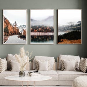 May include: Three framed landscape prints showcasing mountain and forest scenes. The artwork features a road, a lake, and autumnal trees. The colour scheme includes browns, greens, and greys. The prints are displayed above a beige sofa.