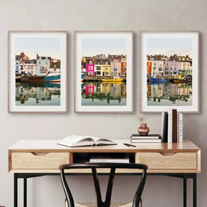 Può includere: Tre stampe artistiche incorniciate con edifici colorati riflessi nell'acqua. Gli edifici sono dipinti in tonalità di blu, rosa, giallo e arancione. Le stampe sono esposte su una scrivania con libri e una sedia.