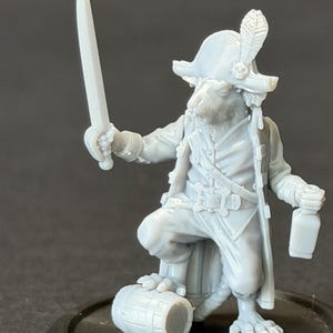 Könnte beinhalten: Miniaturfigur eines Nagetierpiraten, der ein Schwert und eine Flasche hält. Die Figur trägt einen Dreispitz mit Feder, einen langen Mantel und kniehohe Stiefel. Ein kleines Fass steht neben der Figur auf einem runden Sockel.