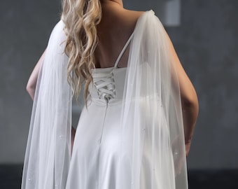 May include: A white floor-length gown featuring a corset-style lace-up back closure and thin shoulder straps. The dress is paired with a sheer, floor-length tulle cape adorned with small, evenly spaced white pearls along the fabric.