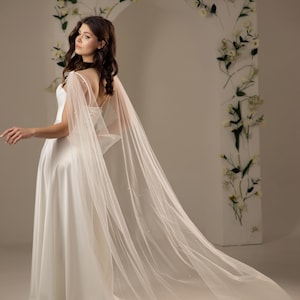 May include: A white wedding dress with a long, flowing tulle cape. The cape is adorned with small, pearl-like embellishments. The bride is standing in front of a white backdrop with a floral arch.