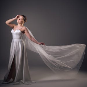 May include: A white satin wedding dress with a high slit and spaghetti straps. A sheer, flowing veil extends from the shoulders. The model is posing with one hand on their head and the other arm extended, creating a sense of movement.