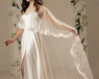 May include: A white satin wedding dress with a lace trim and a sheer cape. The dress has a slit up the front and the cape is flowing in the wind.