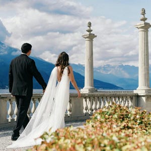 May include: A person in a black suit and a person in a white floor-length gown with a long sheer cape-style veil stand together on a stone terrace with balustrades, overlooking a large lake and distant mountains under a cloudy blue sky.