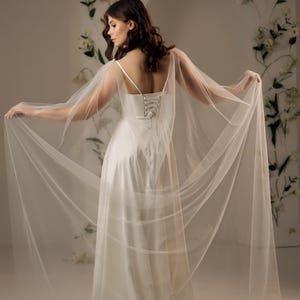 May include: A floor-length white satin gown featuring thin spaghetti straps and a lace-up corset back closure. The dress is complemented by long, sheer, flowing tulle cape sleeves that extend from the shoulders, creating a dramatic, airy silhouette.