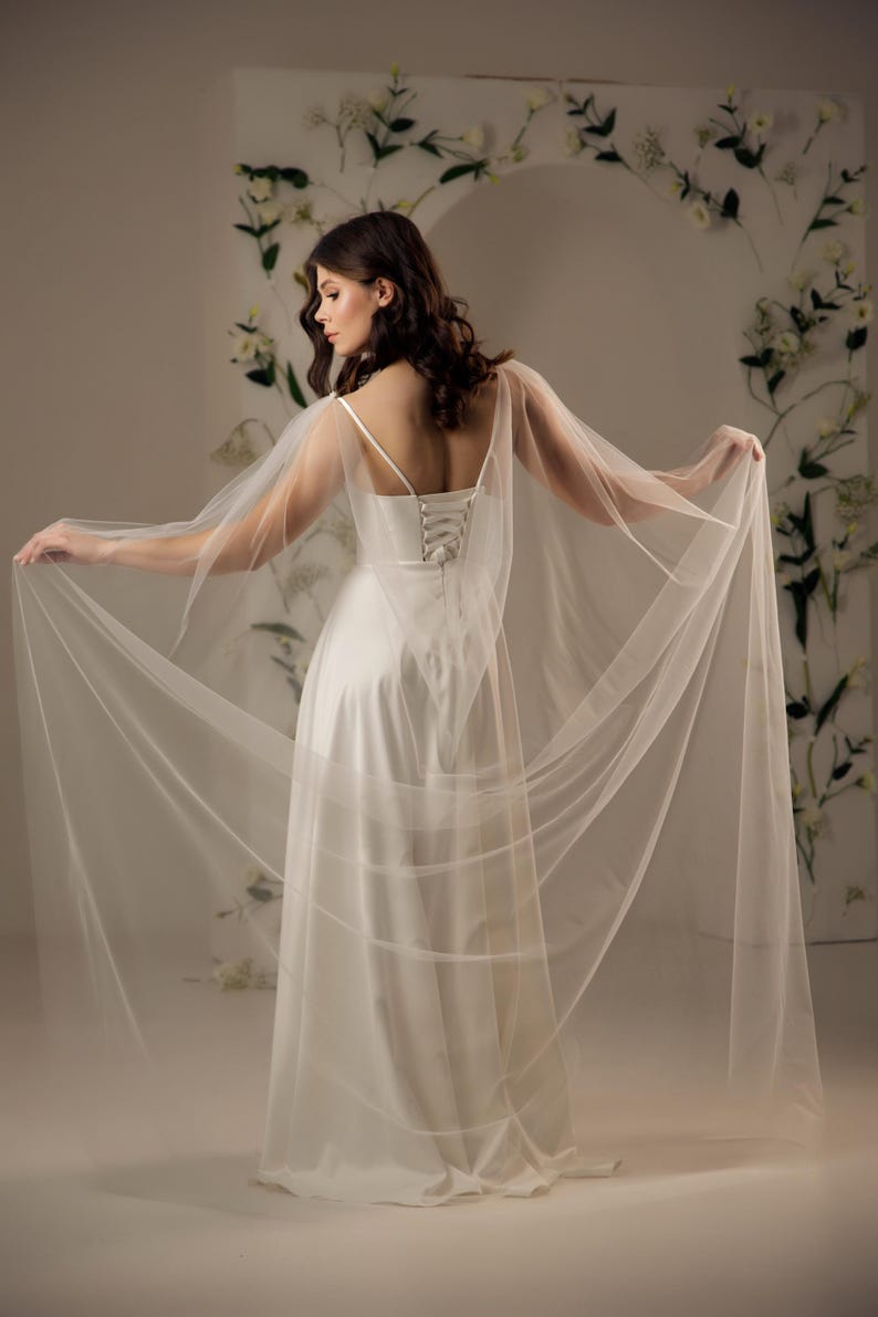 May include: A floor-length white satin gown featuring thin spaghetti straps and a lace-up corset back closure. The dress is complemented by long, sheer, flowing tulle cape sleeves that extend from the shoulders, creating a dramatic, airy silhouette.