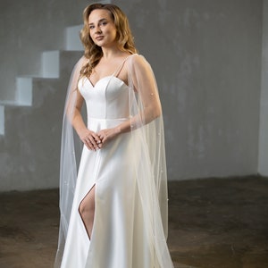 May include: A floor-length white satin dress featuring a sweetheart neckline, thin spaghetti straps, and a high front slit. The dress is paired with a sheer, floor-length tulle cape adorned with small white pearls that drape over the shoulders.