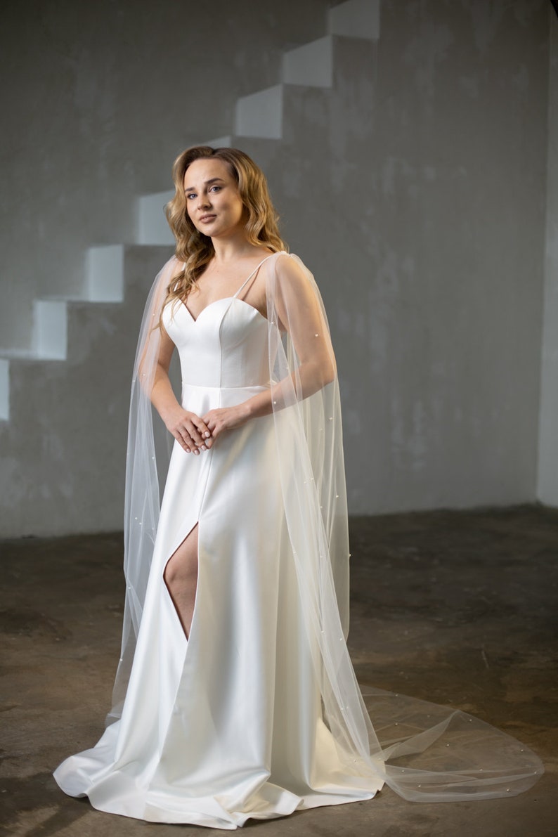May include: A floor-length white satin dress featuring a sweetheart neckline, thin spaghetti straps, and a high front slit. The dress is paired with a sheer, floor-length tulle cape adorned with small white pearls that drape over the shoulders.