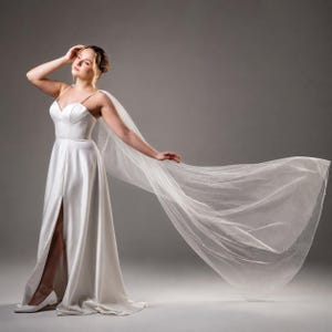 May include: A floor-length white satin gown featuring a sweetheart neckline, thin shoulder straps, and a high leg slit. The dress is paired with a long, sheer, flowing tulle cape that extends outward, creating a dramatic, airy silhouette.