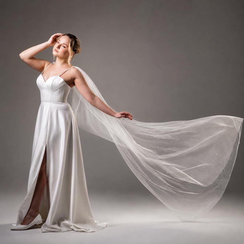 May include: A floor-length white satin gown featuring a sweetheart neckline, thin shoulder straps, and a high leg slit. The dress is paired with a long, sheer, flowing tulle cape that extends outward, creating a dramatic, airy silhouette.