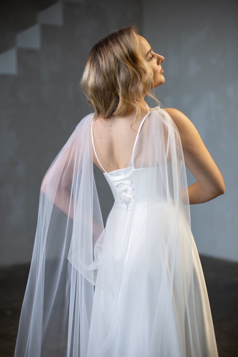 May include: A white floor-length gown featuring thin spaghetti straps, a lace-up corset back closure, and sheer tulle cape sleeves that drape from the shoulders. The bodice is fitted, transitioning into a flowing skirt made of smooth fabric.