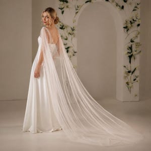 May include: A floor-length white satin gown featuring a sleeveless bodice with thin straps and a long, flowing sheer tulle cape attached at the shoulders that extends behind the garment, creating a dramatic, layered silhouette with a smooth finish.